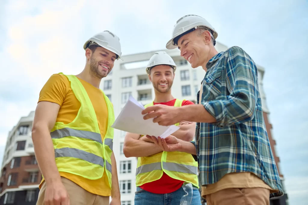 An image of 3 contractor that is smiling and discussing the blueprint of a house.